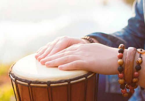 Hands adorned with beaded bracelets are playing a djembe drum in an outdoor setting.