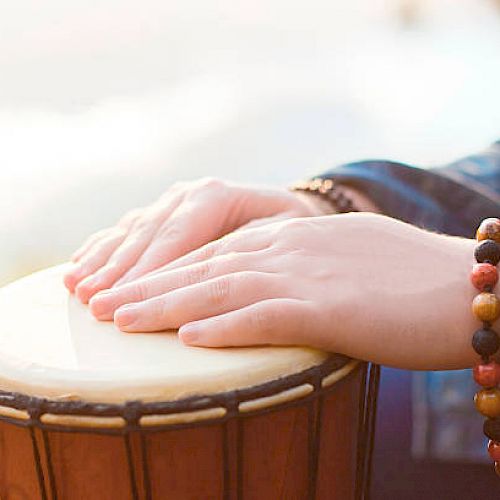 Hands adorned with beaded bracelets are playing a djembe drum in an outdoor setting.