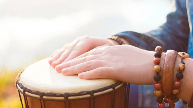 Hands adorned with beaded bracelets are playing a djembe drum in an outdoor setting.