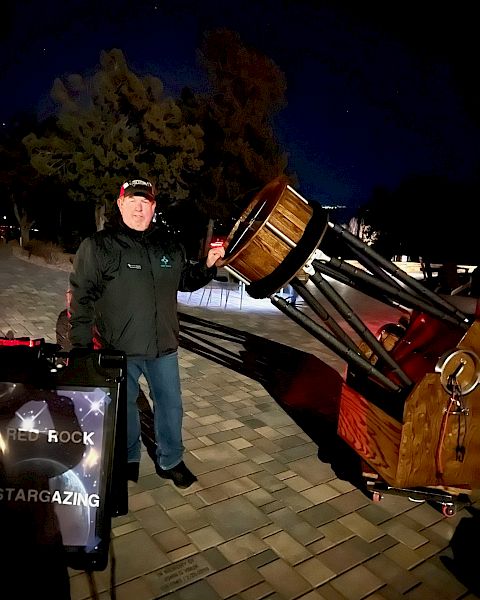 A person stands outside at night beside a large telescope, with a sign that reads 