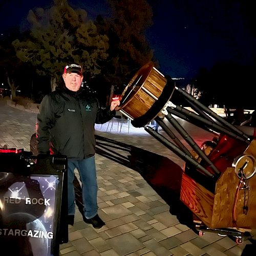 A person stands outside at night beside a large telescope, with a sign that reads 