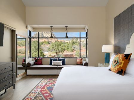 A cozy bedroom with modern decor, large windows showcasing an outdoor view, a flat-screen TV, a dresser, and a window seat.