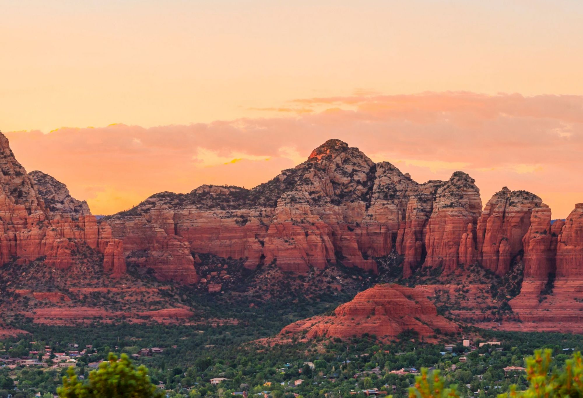 A scenic view of red rock formations and a vibrant sunrise or sunset sky with pink and orange hues, surrounded by vegetation and hints of buildings.