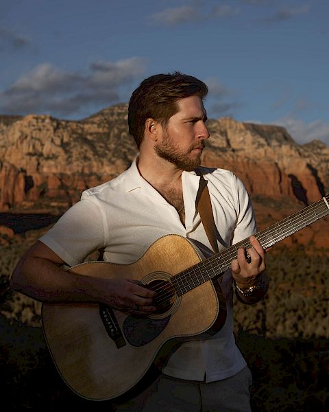 A man playing an acoustic guitar outdoors with a scenic mountain and canyon backdrop, under a partially cloudy sky.