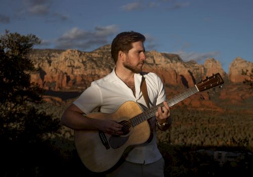 A man playing an acoustic guitar outdoors with a scenic mountain and canyon backdrop, under a partially cloudy sky.