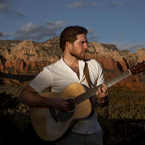 A man playing an acoustic guitar outdoors with a scenic mountain and canyon backdrop, under a partially cloudy sky.