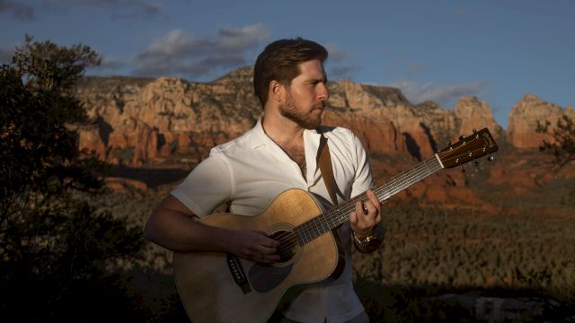 A man playing an acoustic guitar outdoors with a scenic mountain and canyon backdrop, under a partially cloudy sky.