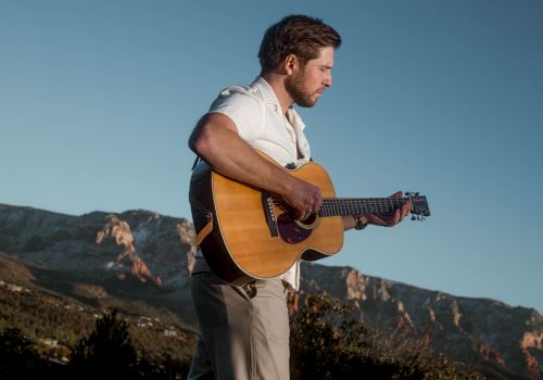 A man in a white shirt is playing an acoustic guitar outdoors, with mountains in the background, under a clear blue sky.