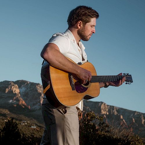 A man in a white shirt is playing an acoustic guitar outdoors, with mountains in the background, under a clear blue sky.