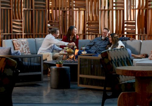 Four people are seated around a fire pit in a cozy setting with wooden decor, enjoying food and drinks, and engaging in conversation.