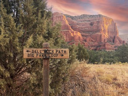 A wooden signpost directs to Bell Rock Path and Big Park Loop, set against a backdrop of red rock mountains and a colorful sky at sunset.