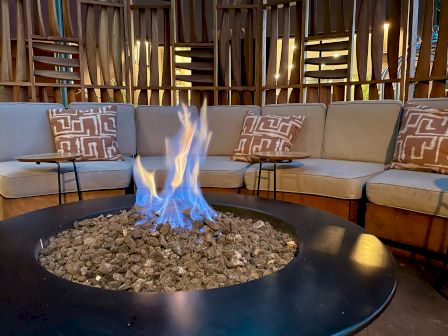 A cozy seating area with a circular fire pit in the center, surrounded by cushions and wooden decor.