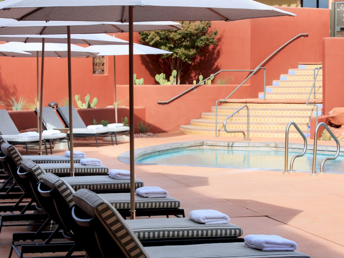 The image shows a serene pool area with lounge chairs, each adorned with a rolled towel. There are umbrellas providing shade and stairs nearby.