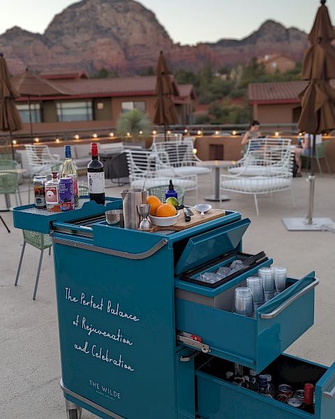 An outdoor seating area with a mountain view features a blue bar cart filled with various bottles and tools for making drinks.