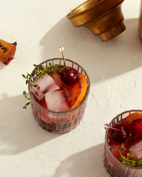 Two cocktail glasses with ice, garnished with cherries and herbs, an empty brass container, and a wooden muddler on a light surface end the sentence.