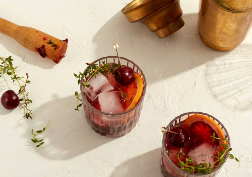 Two cocktail glasses with ice, garnished with cherries and herbs, an empty brass container, and a wooden muddler on a light surface end the sentence.