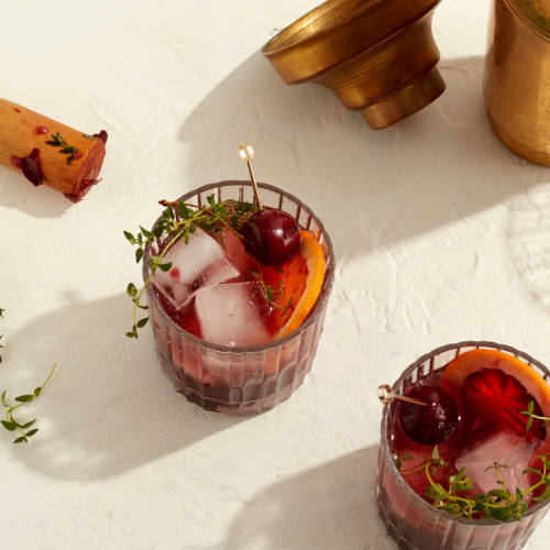 Two cocktail glasses with ice, garnished with cherries and herbs, an empty brass container, and a wooden muddler on a light surface end the sentence.