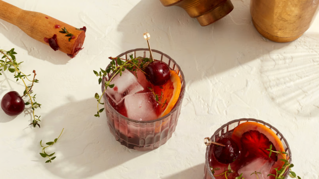 Two cocktail glasses with ice, garnished with cherries and herbs, an empty brass container, and a wooden muddler on a light surface end the sentence.