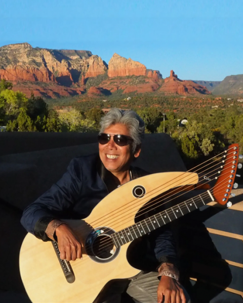 A person with sunglasses holds a harp guitar, sitting outdoors with a stunning backdrop of red rock formations and blue sky.