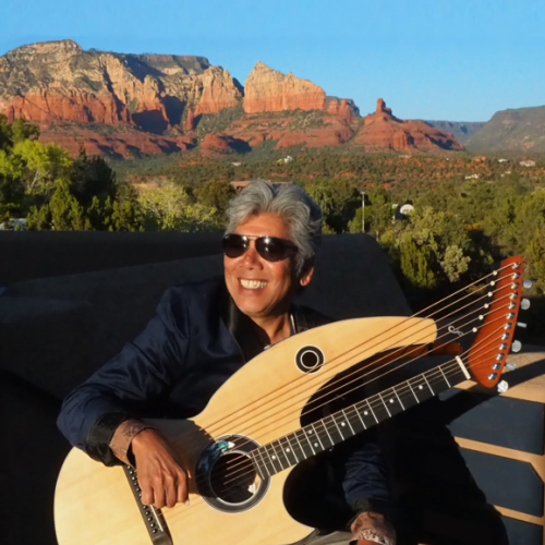 A person with sunglasses holds a harp guitar, sitting outdoors with a stunning backdrop of red rock formations and blue sky.