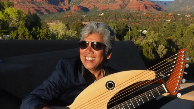 A person with sunglasses holds a harp guitar, sitting outdoors with a stunning backdrop of red rock formations and blue sky.