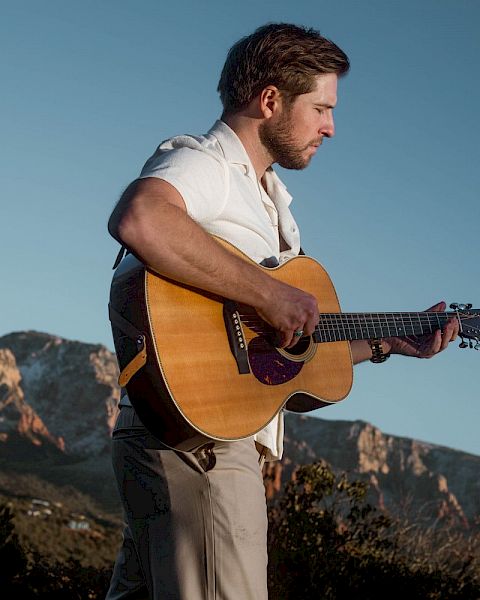 A man is playing a guitar outdoors, with a mountainous landscape in the background under a clear blue sky.