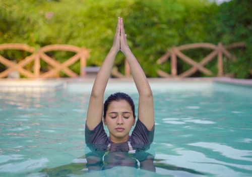 A person is in a pool, eyes closed, with hands raised together above their head, surrounded by greenery and wooden fencing.