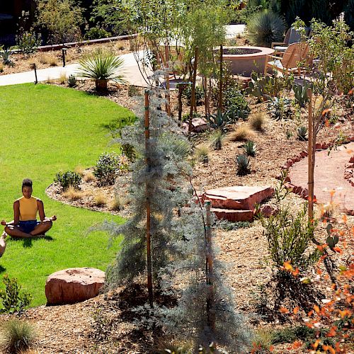 Two people are meditating on a grassy lawn in an outdoor garden surrounded by trees and shrubs. There is a circular pathway and rocks nearby.