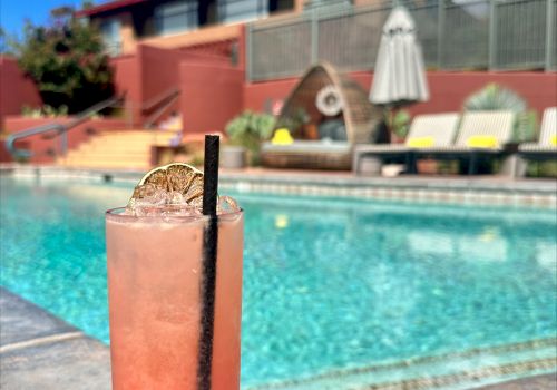 A tall pink drink with a straw sits by a pool on a sunny day, with lounge chairs and a building in the background.