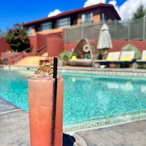 A tall pink drink with a straw sits by a pool on a sunny day, with lounge chairs and a building in the background.