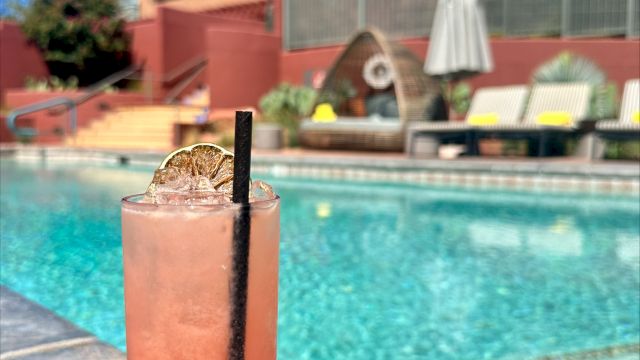 A tall pink drink with a straw sits by a pool on a sunny day, with lounge chairs and a building in the background.