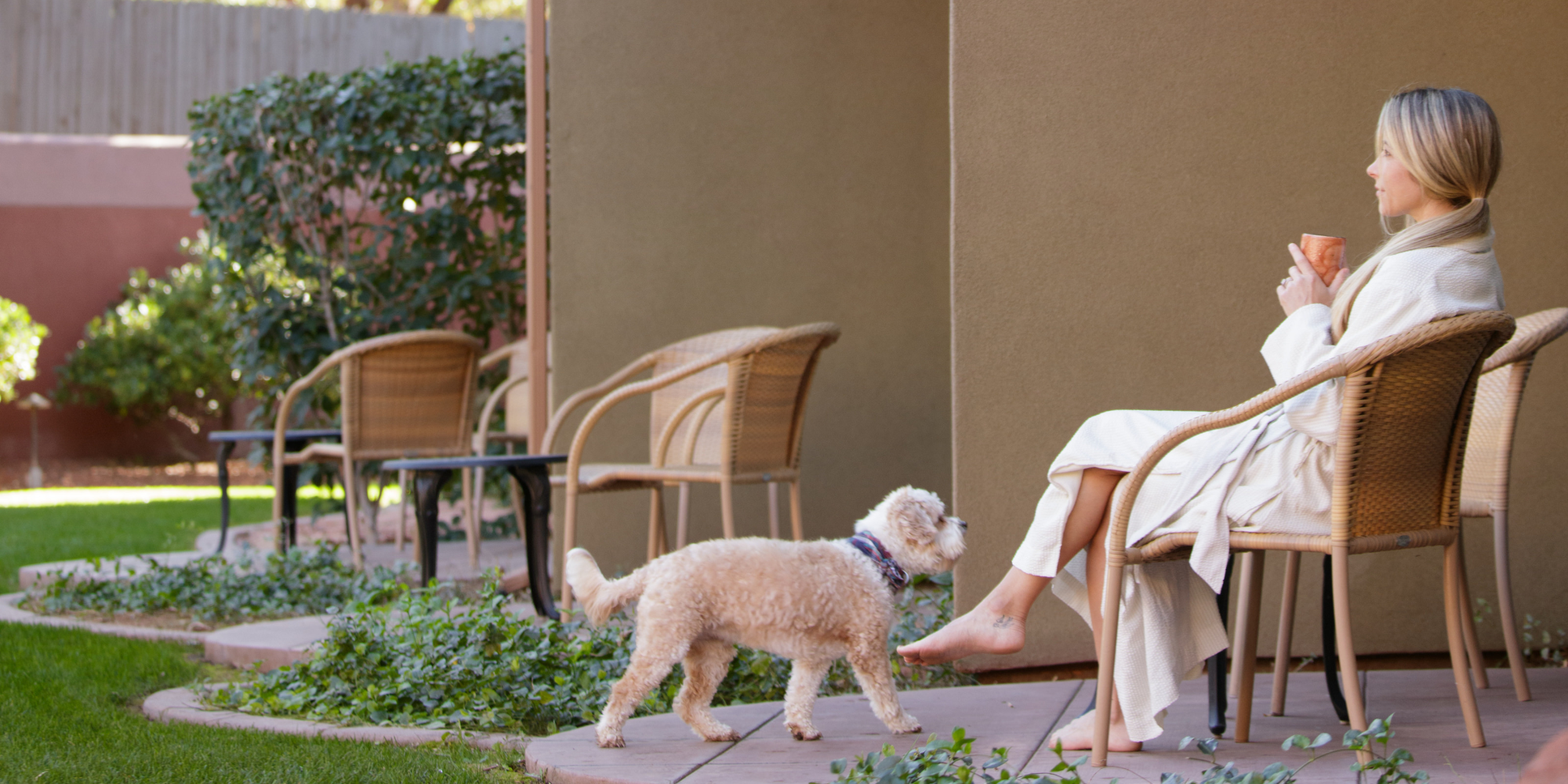 A person sits, enjoying a beverage on a patio chair while wearing a bathrobe, and a small dog walks nearby in a garden setting.