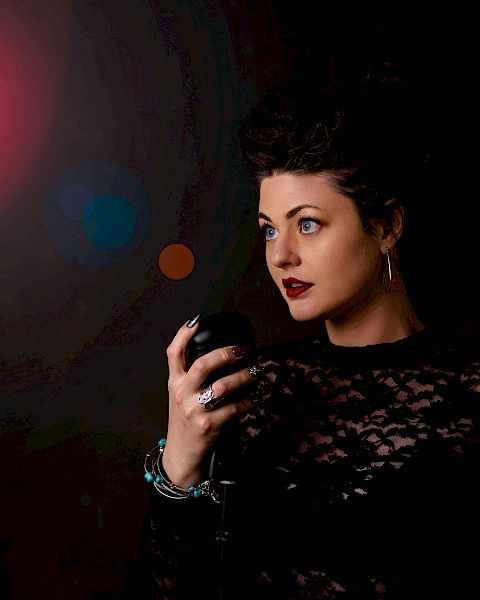 A woman in a black lace outfit with dark red lipstick stands with her hand raised against a dark background illuminated by a red light.