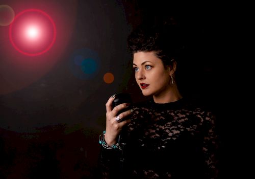 A woman in a black lace outfit with dark red lipstick stands with her hand raised against a dark background illuminated by a red light.