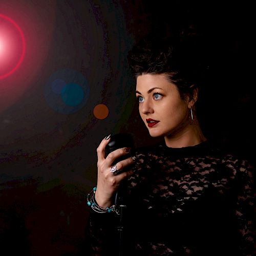 A woman in a black lace outfit with dark red lipstick stands with her hand raised against a dark background illuminated by a red light.