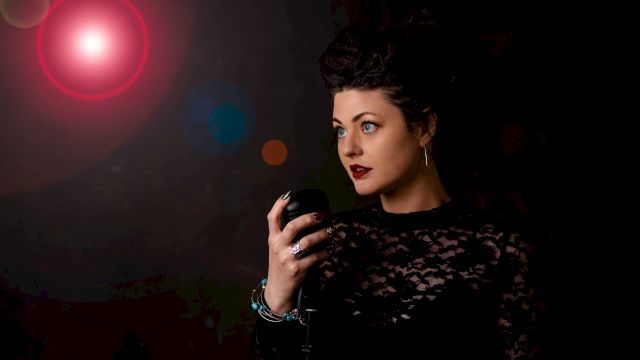 A woman in a black lace outfit with dark red lipstick stands with her hand raised against a dark background illuminated by a red light.