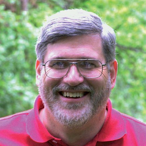 A smiling person with glasses, gray hair, and a beard is wearing a red shirt and standing in front of a green, outdoor background.