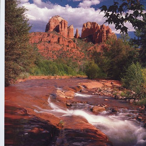 A scenic landscape features a flowing river with red rock formations and greenery under a blue sky with clouds in the background.