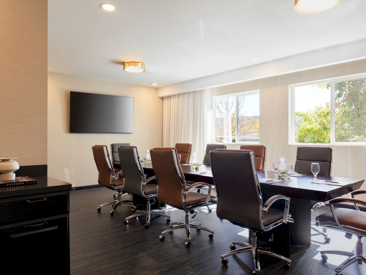 A modern conference room with a large table, chairs, a wall-mounted TV, and windows allowing natural light to come in.