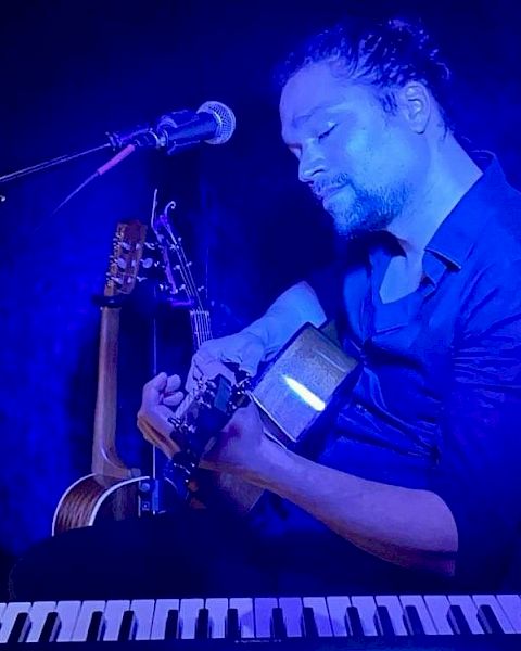 A person is playing a guitar with a microphone nearby, additional guitars in the background, and a keyboard visible at the bottom of the image.