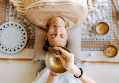 A person is lying down with a singing bowl being used above them. Other bowls and a patterned cloth are visible around the person.