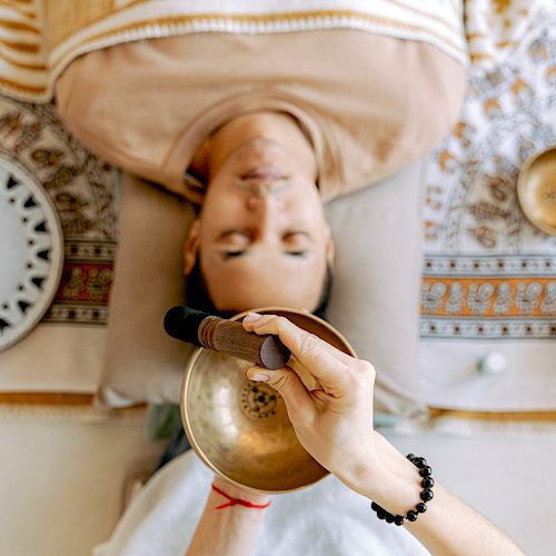 A person is lying down with a singing bowl being used above them. Other bowls and a patterned cloth are visible around the person.