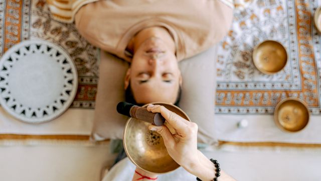 A person is lying down with a singing bowl being used above them. Other bowls and a patterned cloth are visible around the person.