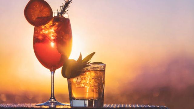 Two cocktails with ice and citrus garnishes, set against a vibrant sunset background.