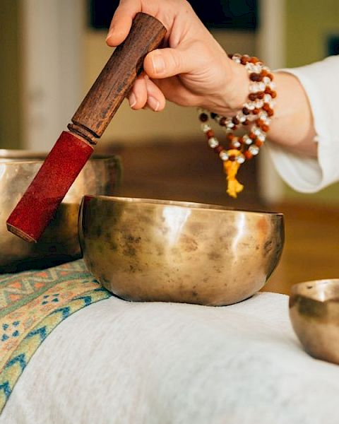 A person is using a mallet to play Tibetan singing bowls placed on a cloth-covered surface, likely for a sound healing session.