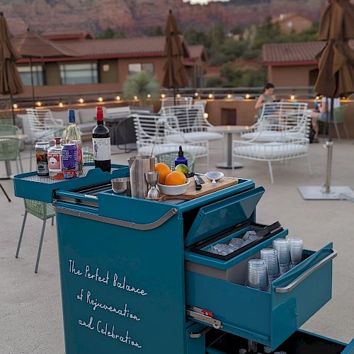 An outdoor seating area with a mountain view features a blue bar cart filled with various bottles and tools for making drinks.