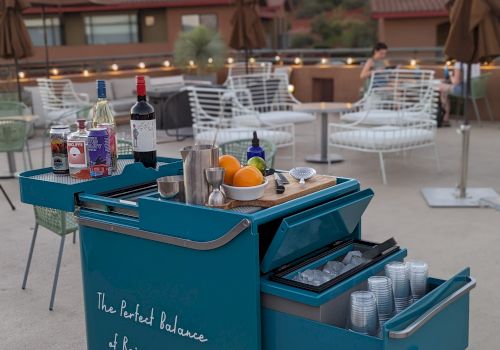 An outdoor seating area with a mountain view features a blue bar cart filled with various bottles and tools for making drinks.