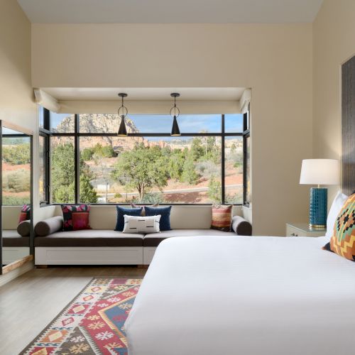 A cozy bedroom with a large bed, patterned rug, and a seating area with a view of the outdoors through large windows.