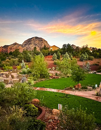 A landscaped garden with lush greenery and a pathway, set against a mountain backdrop and a colorful sunset sky.