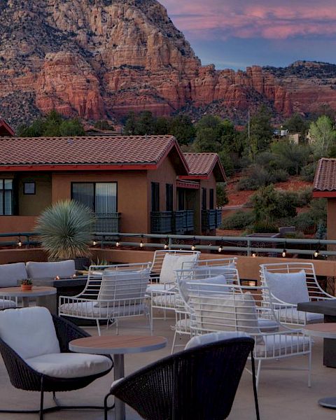 A rooftop patio with seating and fire pits, surrounded by buildings and stunning red rock mountains in the background, under a pinkish sky.